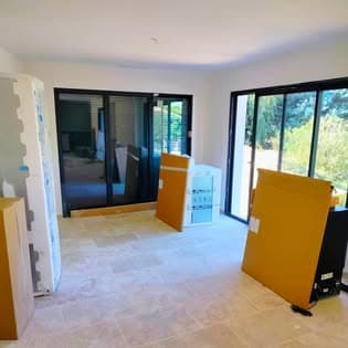 Rénovation intérieure : baies vitrées coulissantes à cadre noir, sol en dalles travertin beige et murs blancs, lumière naturelle sur jardin