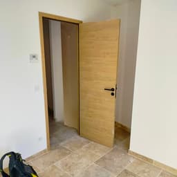 Porte intérieure bois clair avec poignée et rosace noires, encadrement assorti, sol travertin et plinthes pierre