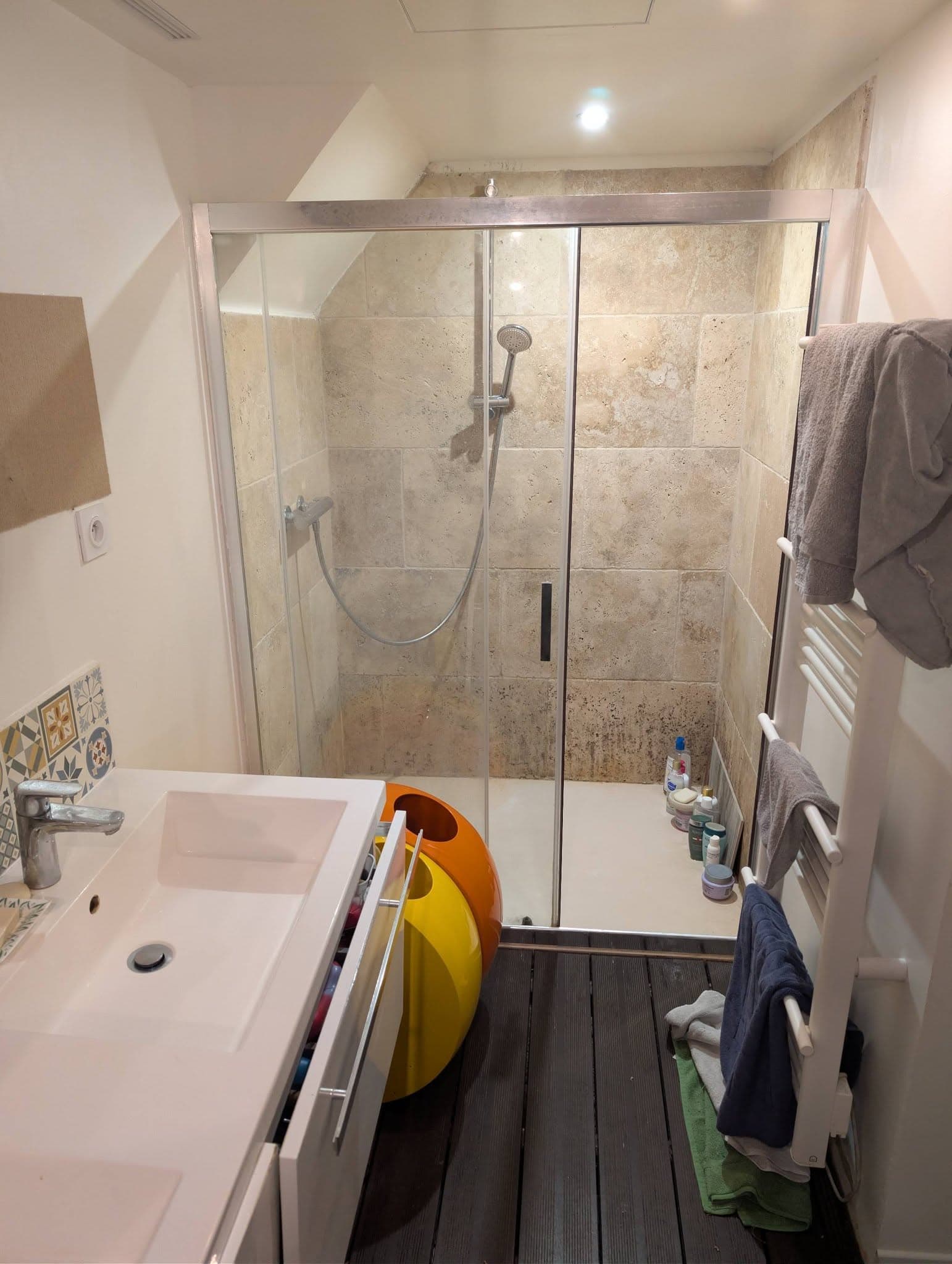 Vue frontale de salle d'eau avec cabine de douche vitrée, murs effet pierre et meuble vasque blanc